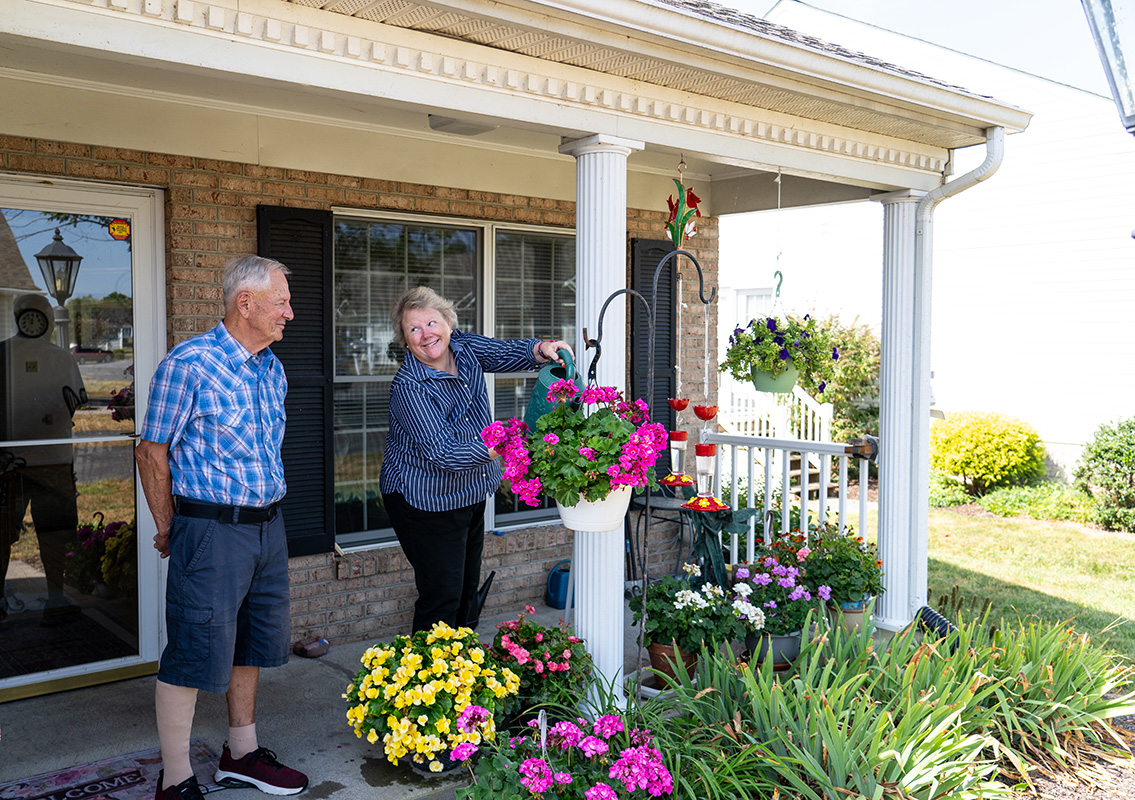 An elderly couple standing outside their residence watering their flowerbed